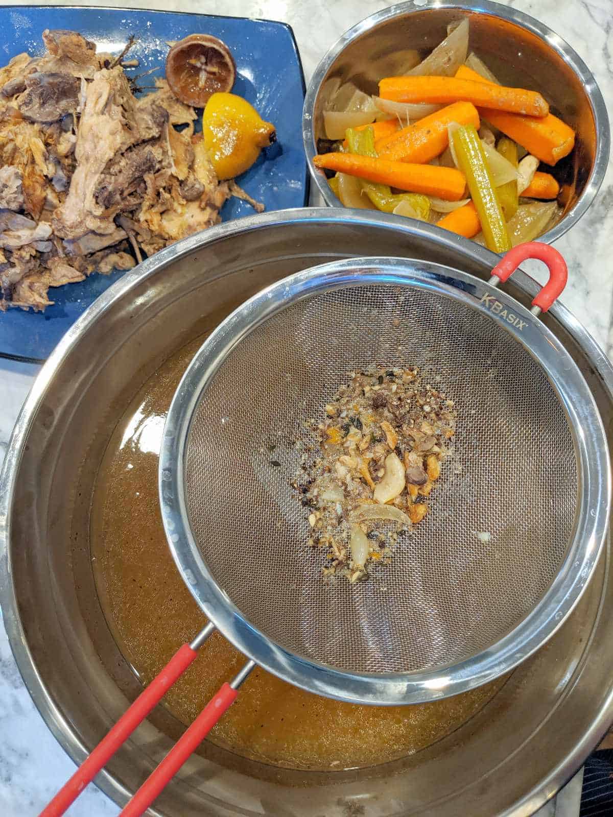 Overhead view of strainer setting on a large bowl of broth with recently removed bones and vegetables in the background.