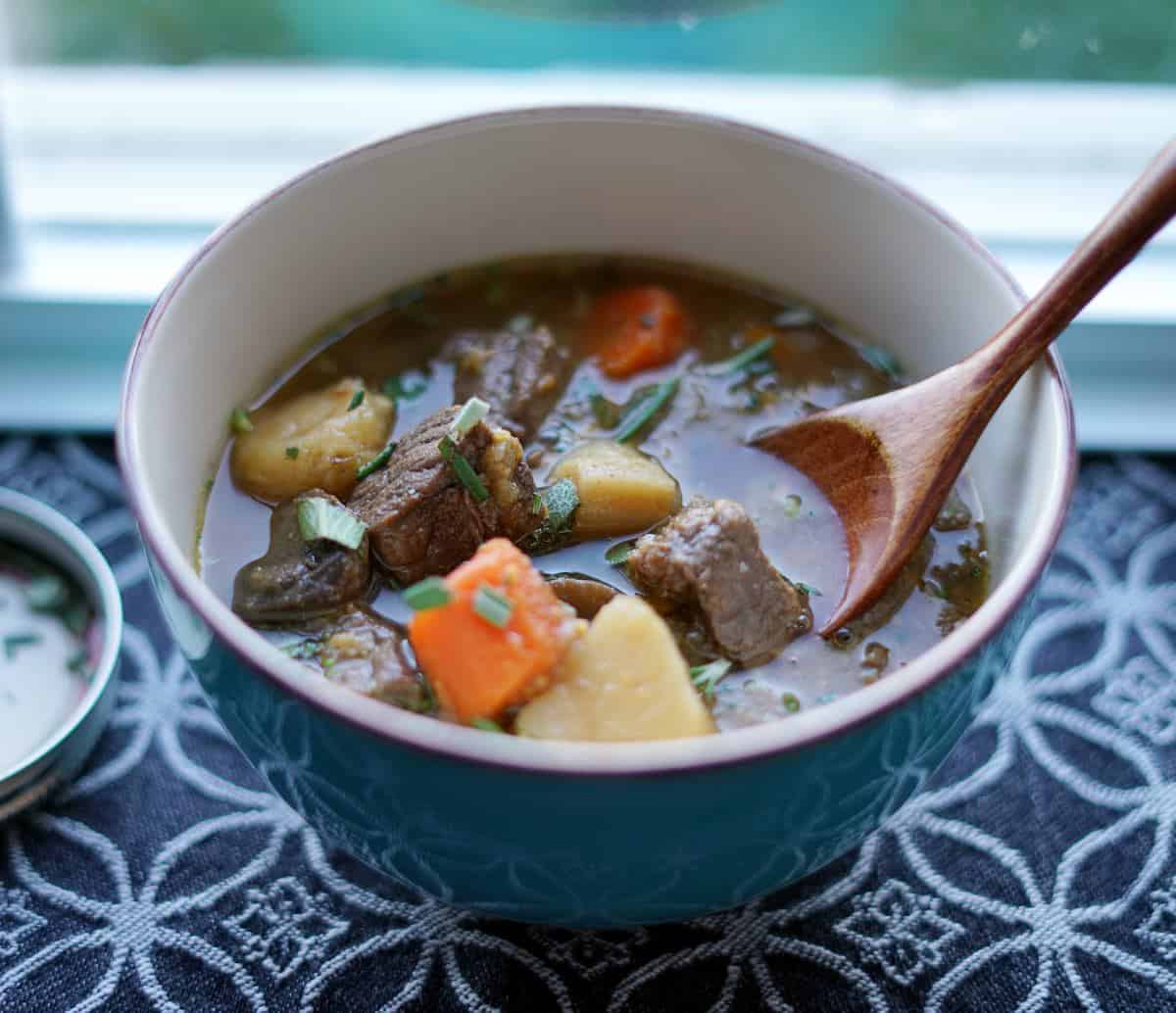 Bowl of beef stew with a wooden spoon in it, sitting next to a window.