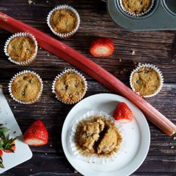 Several strawberry rhubarm muffins on a table with one of them on a plate near a piece of rhubarb.