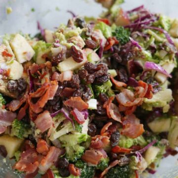 Close up of broccoli salad in a glass bowl with raisins, bacon, and red onions and two wooden salad spoons.