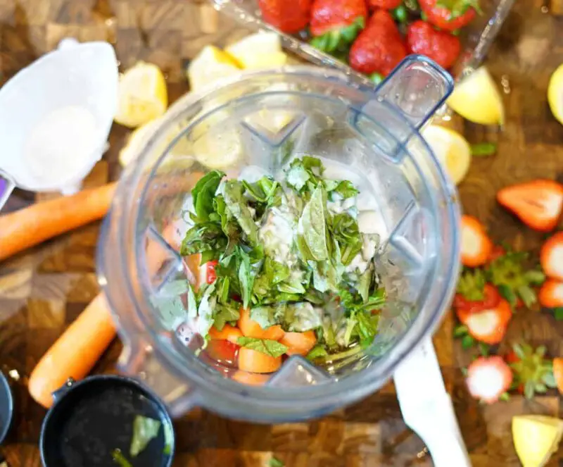 basil, carrots, strawberries, and coconut milk in a blender with various fruit on a cutting board in the background