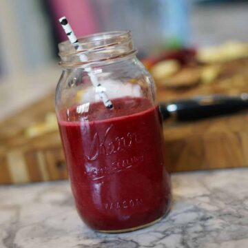 Red smoothie in a mason jar with a wooden cutting board and fruit scraps in the background.