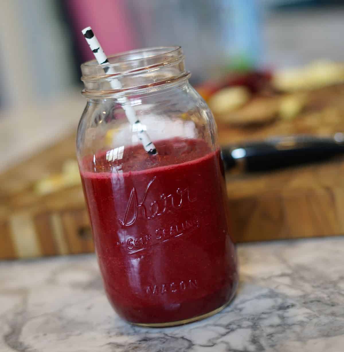 Red smoothie in a mason jar with a wooden cutting board and fruit scraps in the background.