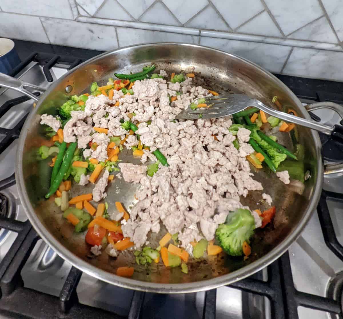 Cooked ground chicken on top of stir fry vegetables in a large metal pan.