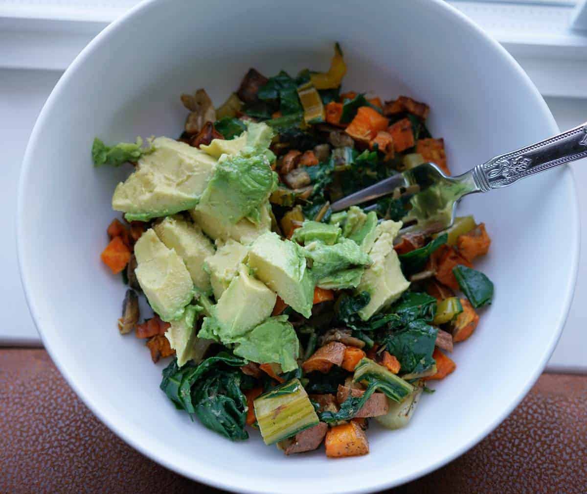 Rainbow chard, sweet potatoes, and mushrooms in a white bowl with avocado on top.