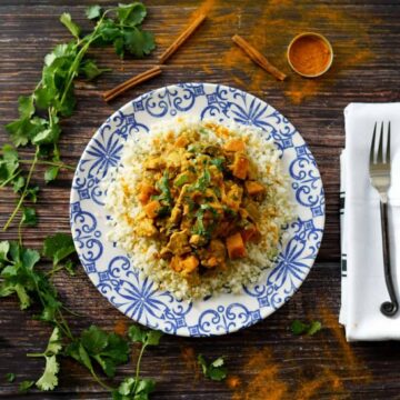 AIP moroccan chicken stew on a blue and white plate with silverware on a napkin and spice and herbs sprinkled on the table