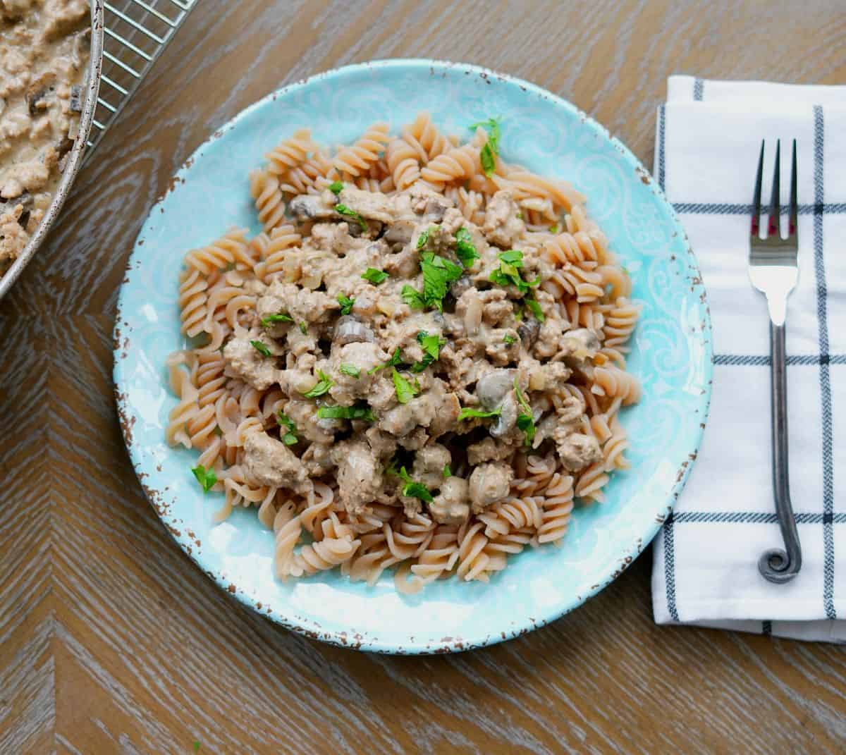 Ground turkey stroganoff over a bed of noddles on a blue plate next to a napkin and fork.