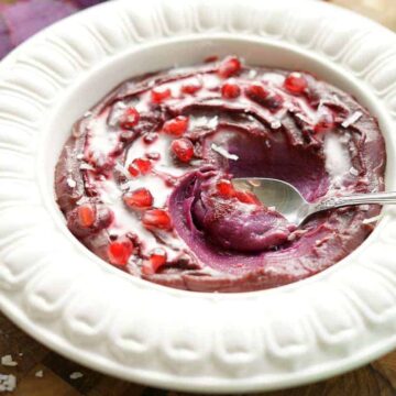 puple sweet potato bowl topped with pomegranate arils and coconut milk with a spoon in the middle.