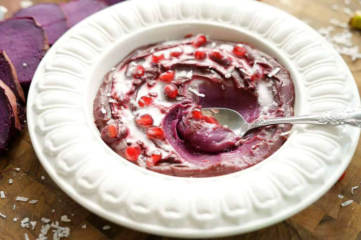 puple sweet potato bowl topped with pomegranate arils and coconut milk with a spoon in the middle.