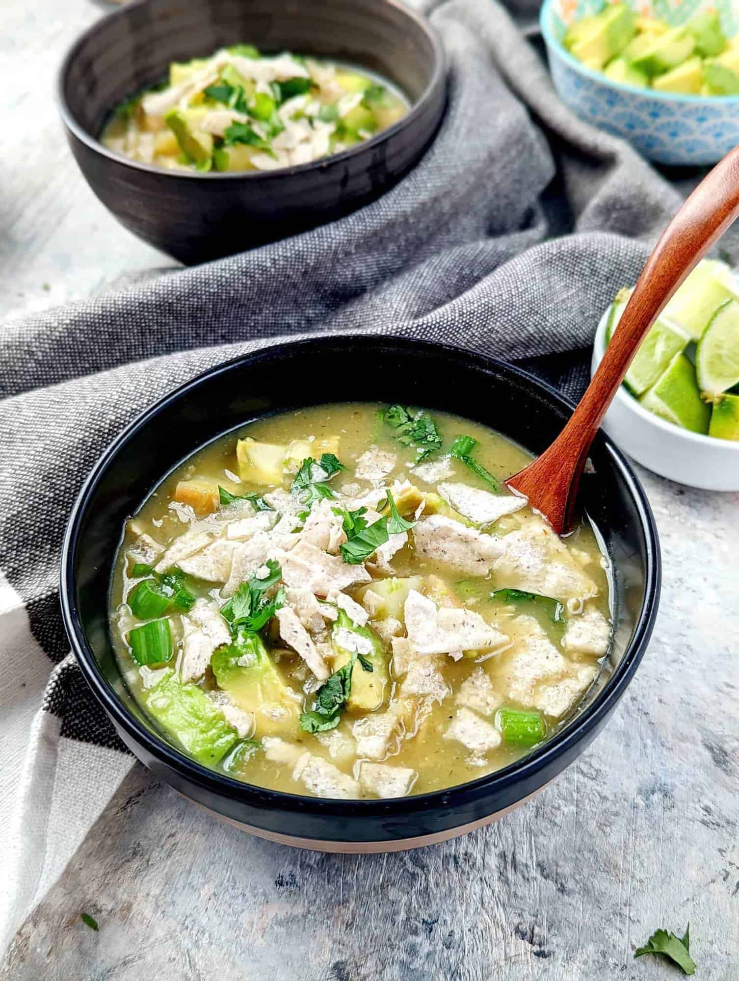nightshade free green chicken enchilada soup in 2 black bowls with wooden spoons and bowls of avocados and limes in the background.