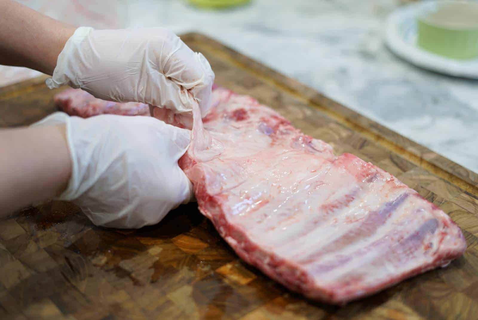 gloved hands peeling the silverskin off the back of raw pork ribs