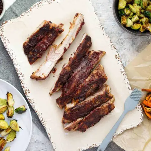 air fryer pork ribs with bbq sauce on a tan platter surrounded by sweet potato fries, brussels sprouts, a dish of bbq sauce, and a plate of ribs and veggies.