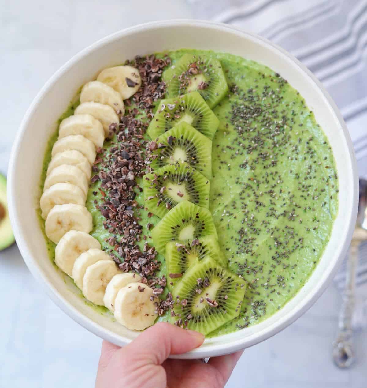 Overhead view of avocado banana kiwi smoothie bowl with sliced bananas, kiwis, cacao nibs, and chia seeds.