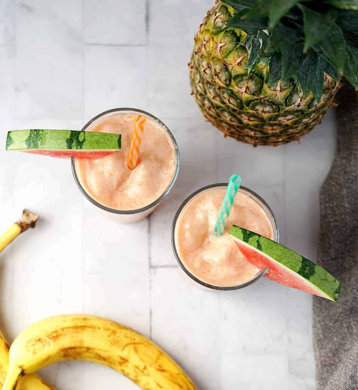 Overhead view of two glasses of watermelon smoothie with straws and watermelon slices and whole pineapple in the background.