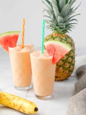 Two glasses of watermelon smoothie with straws and watermelon slices and whole pineapple in the background.