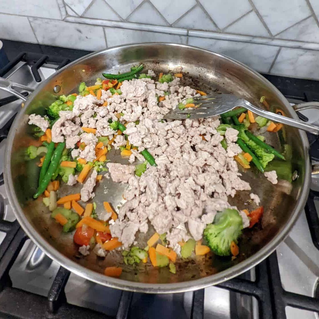 Cooked ground chicken on top of stir fry vegetables in a large metal pan.